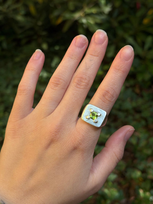 A close-up of a hand wearing a square silver ring with a central green gemstone, set against a blurred green outdoor background.